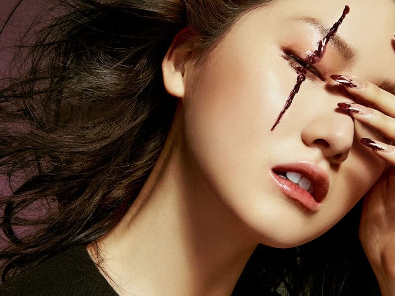 A woman with dramatic makeup and a painted face, featuring a cross design over her eye.